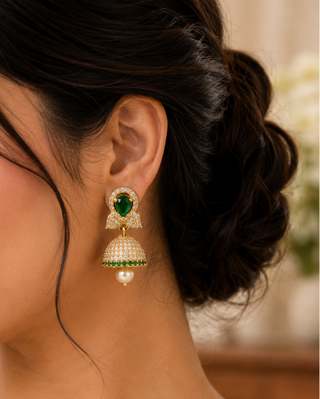 A woman wearing a cream-colored saree and matching cream jewelry, featuring a prominent gold-toned jhumka earring with a green stone and pearl drop.