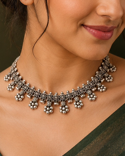 A woman wearing a dark green saree and a silver-toned traditional choker necklace with floral motifs, set against a dark, warm-toned background.