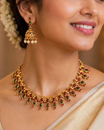 A woman wearing a gold-toned traditional necklace with green stone accents, matching drop earrings, and a cream-colored saree, shown in a warm, softly lit indoor setting.