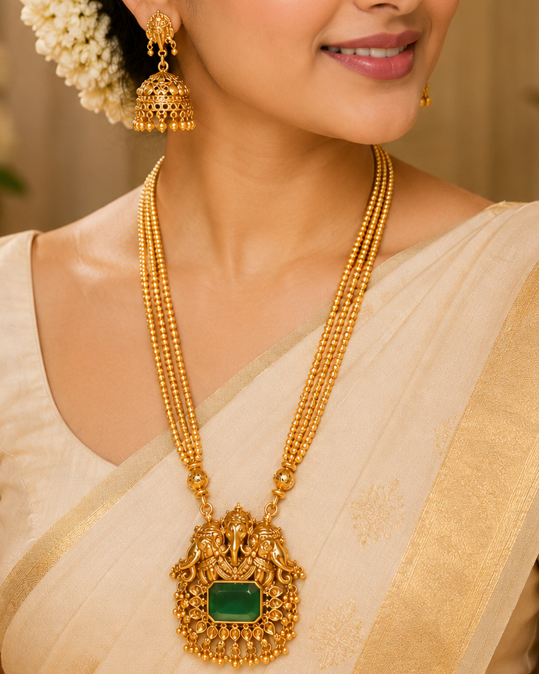 A woman wearing an ornate gold necklace with a large green stone pendant and matching gold earrings, styled with a cream sari and white floral hair accessory.