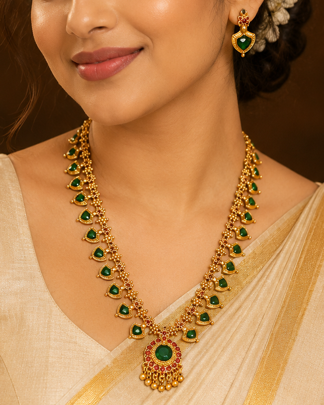A woman wearing a cream-colored saree with gold borders and a matching gold and green jewelry set, including a necklace and earrings, in a warm, elegant indoor setting.