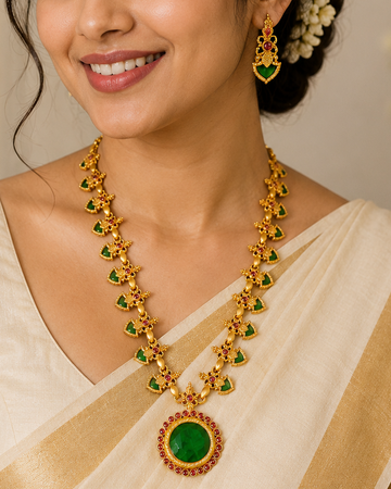 A woman wearing a traditional Indian outfit with a cream and gold saree, accessorized with a gold and green necklace and matching earrings.