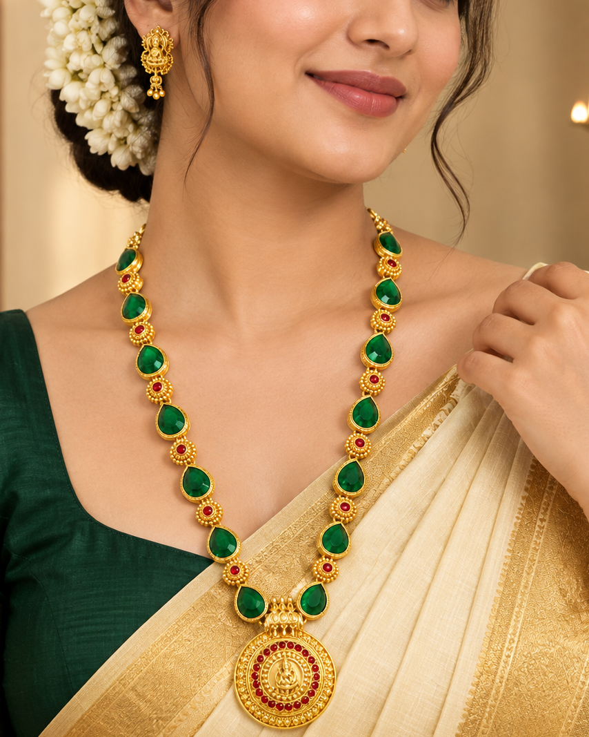 A woman wearing a traditional Indian outfit with a cream and gold saree and a dark green blouse, accessorized with a gold necklace featuring green stones and a matching gold earring.