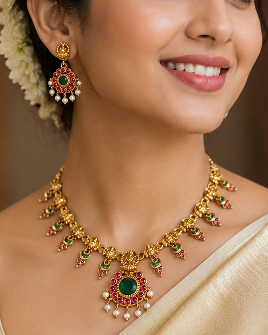 A woman wearing a gold-toned traditional necklace with a green and red pendant, matching earrings, and a cream-colored saree, shown in a warm indoor setting.