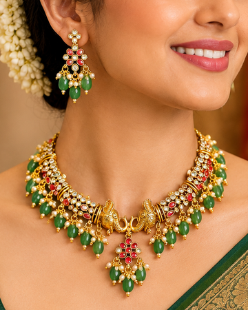 A woman wearing a dark green saree with a gold border and a matching gold, green, and red statement necklace and earrings, posed against a traditional red and gold backdrop.