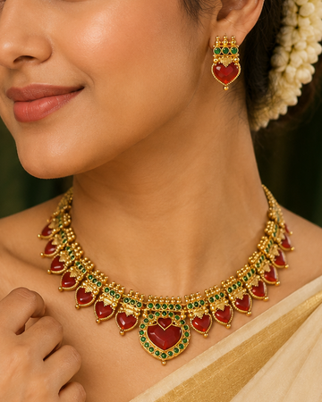A woman wearing a gold heart-shaped necklace and matching heart-shaped earrings with red and green stone accents, styled with a cream and gold saree.