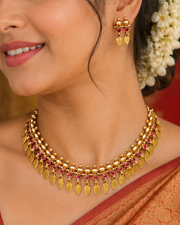 A woman wearing a gold-toned traditional necklace with red stones and yellow teardrop pendants, styled with a red and gold saree and white floral hair accessory.