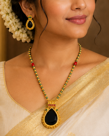 A woman wearing a cream and gold saree with a gold necklace featuring a black teardrop pendant and matching gold earrings, styled with white flowers in her hair.