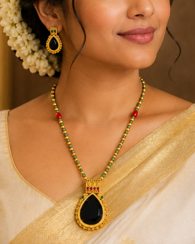 A woman wearing a cream and gold saree with a gold necklace featuring a black teardrop pendant and matching gold earrings, styled with white flowers in her hair.
