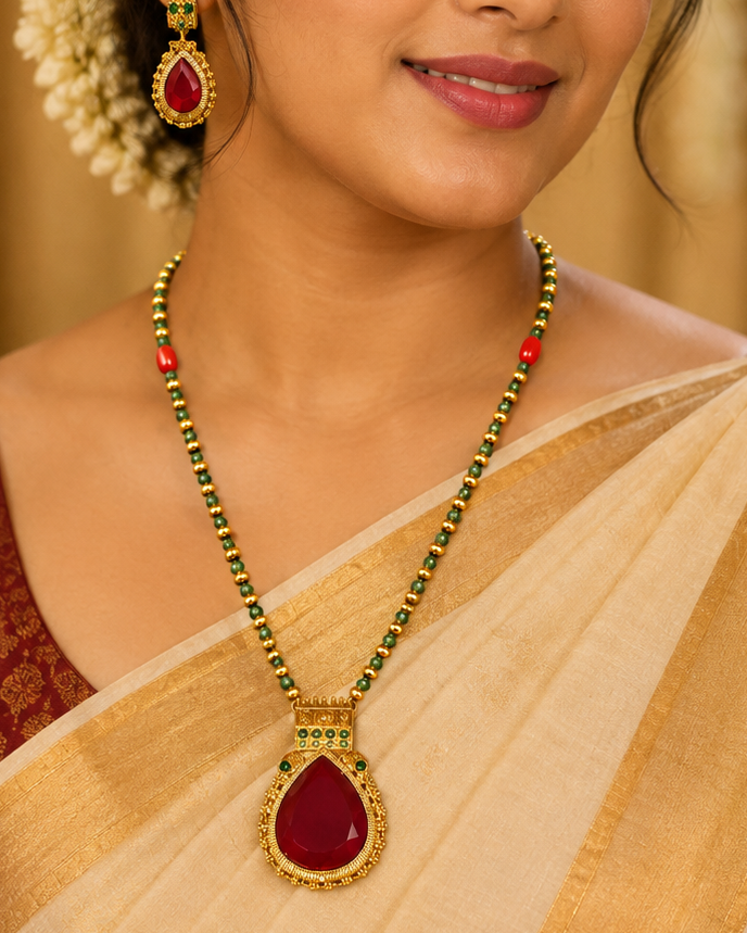 A woman wearing a traditional Indian outfit with a cream and gold saree, a maroon blouse, and a gold necklace with a large red teardrop pendant, along with matching earrings.