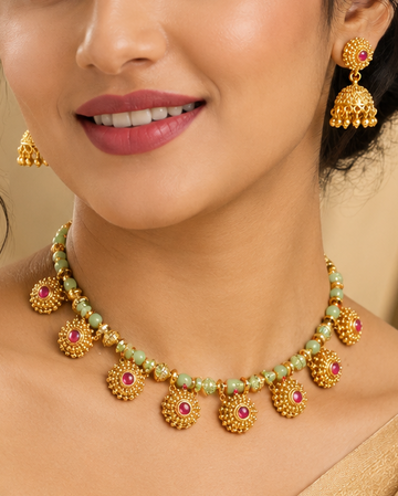 A woman wearing a gold-toned traditional necklace with green and pink stone accents and matching dangling earrings, paired with a beige sari against a soft beige background.