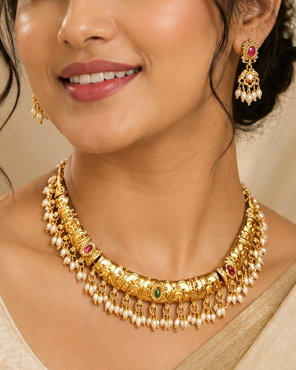 A woman wearing a gold-toned traditional necklace with red and green stone accents, matching earrings, and a cream-colored sari, shown in a softly lit indoor setting.