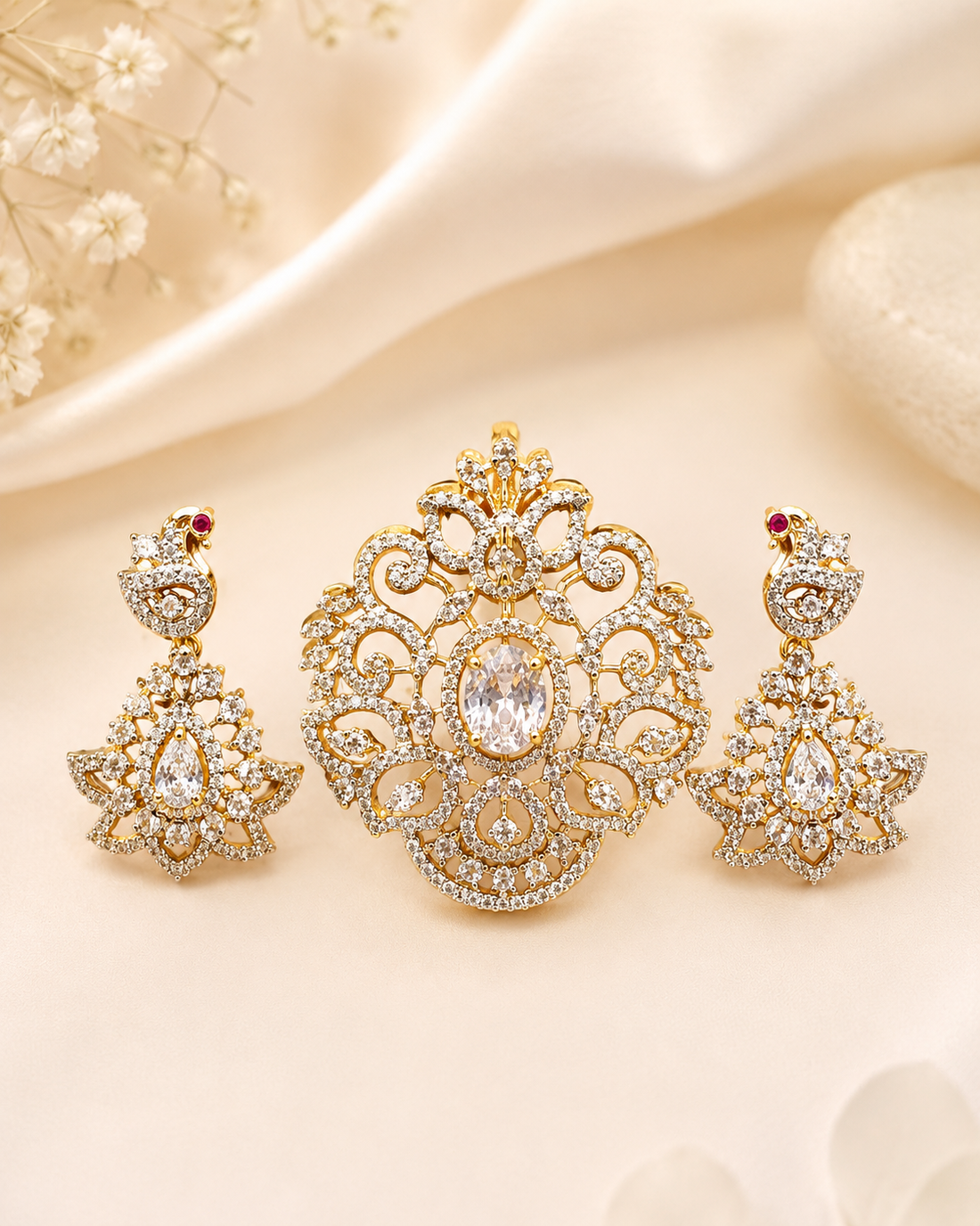 A woman wearing an ornate gold pendant necklace and matching drop earrings with clear stones, styled with a cream-colored sari in a softly lit indoor setting.