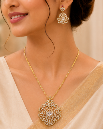 A woman wearing an ornate gold pendant necklace and matching drop earrings with clear stones, styled with a cream-colored sari in a softly lit indoor setting.