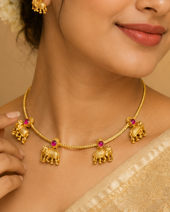 A woman wearing a gold-toned traditional necklace with elephant-shaped charms and matching earrings, styled with a cream-colored saree in a warm, elegant indoor setting.