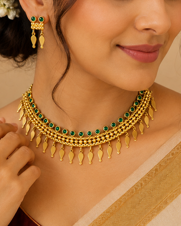 A woman wearing a traditional Indian outfit with a cream and maroon saree, accessorized with a gold choker necklace with green stones, matching earrings, and a gold bangle.