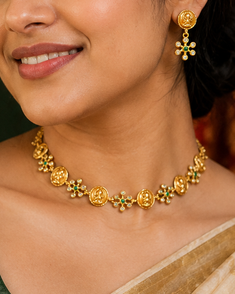 A woman wearing a dark green blouse and a cream saree with gold zari borders, styled with a gold floral necklace and matching earrings, set against a dark green and red patterned backdrop.