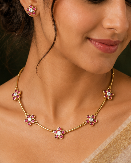 A woman wearing a gold-toned necklace and matching stud earring, each decorated with pink gemstones, styled with a beige sari against a dark green and gold background.