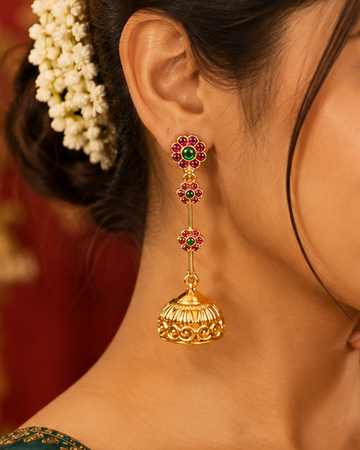 A woman wearing ornate gold jhumka earrings with red and green stone accents, styled with a white floral hair accessory and a dark green blouse with gold embroidery, set against a warm red background.