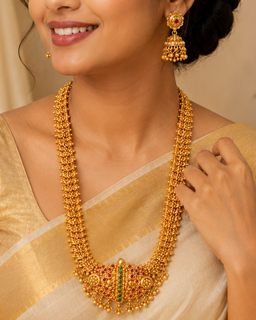 A woman wearing a traditional Indian saree and gold jewelry, featuring a gold-toned necklace with a central green stone accent, matching earrings, and a bracelet.