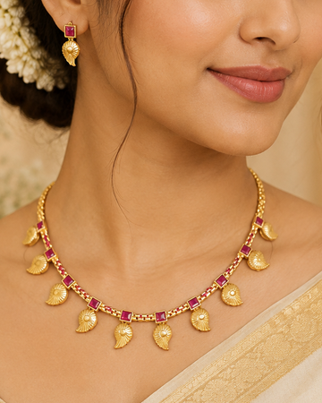 A woman wearing a gold necklace with small red gemstone accents and matching gold earrings, styled with a cream-colored sari against a soft beige background.
