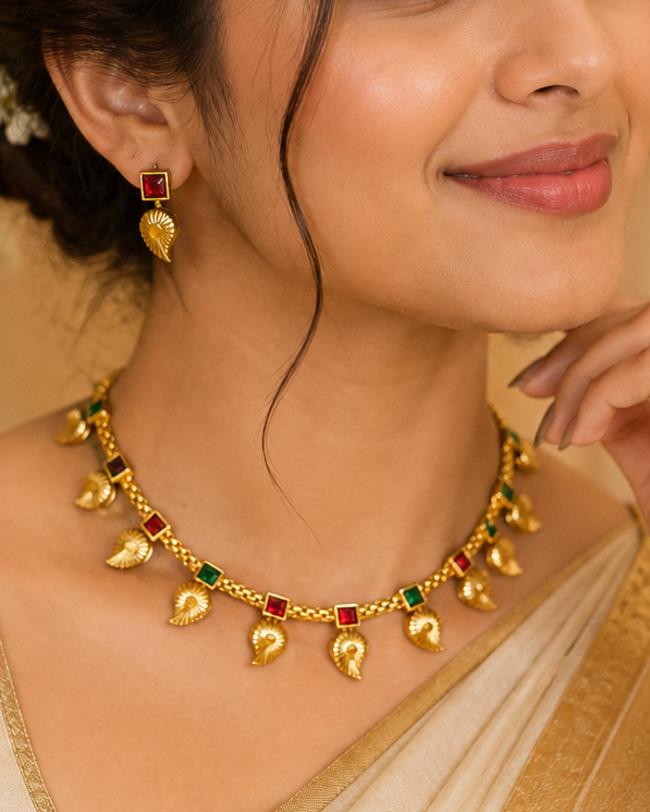 A woman wearing a gold-toned traditional necklace with small leaf-shaped charms and red and green stone accents, paired with matching earrings, in a warm beige saree.