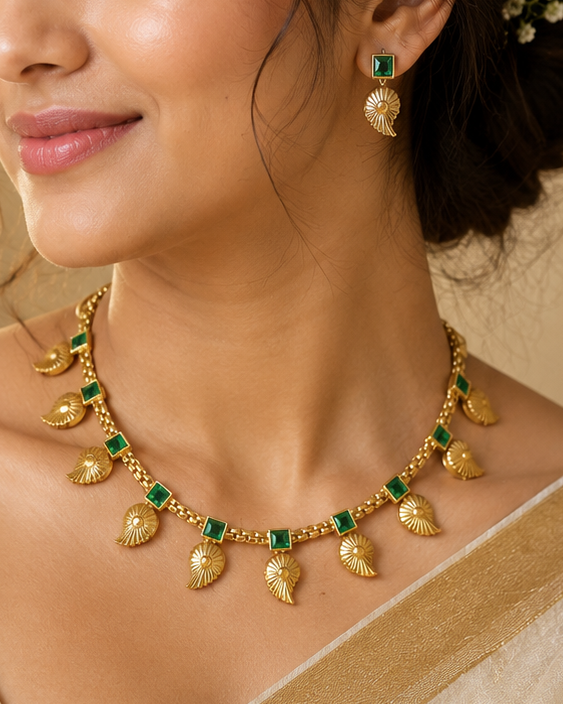 A woman wearing a gold-toned traditional necklace with green stone accents and matching drop earrings, styled with a beige saree in a softly lit indoor setting.