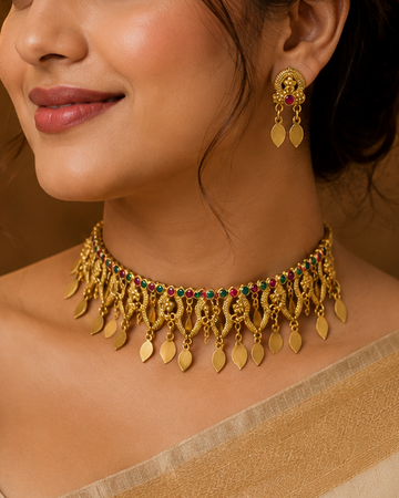 A woman wearing an ornate gold-toned traditional necklace with dangling leaf-shaped charms and matching earrings, styled with a beige saree in a warm, elegant indoor setting.