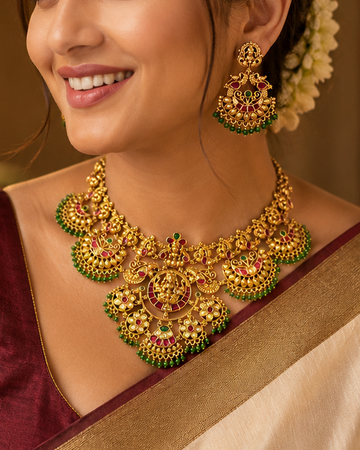 A woman wearing a traditional Indian outfit with a cream and maroon saree, styled with an ornate gold necklace, matching earrings, and a gold bracelet, set against a warm, softly blurred background.