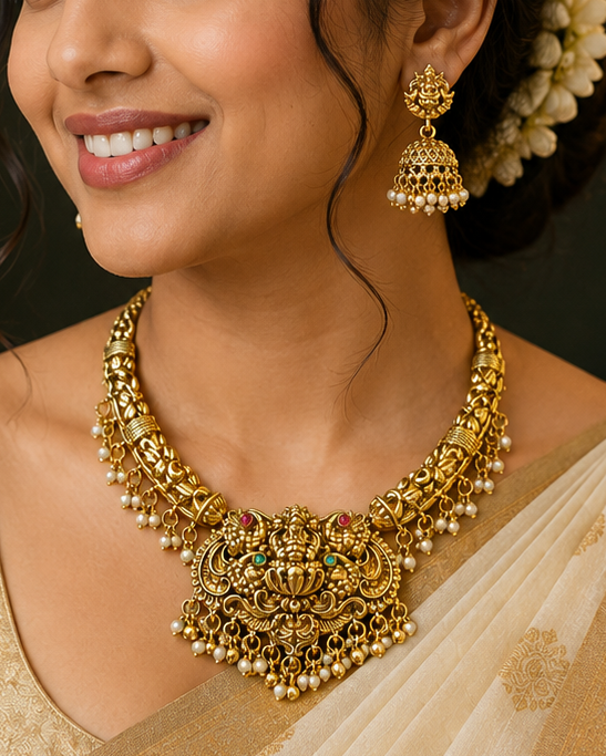 A woman wearing an ornate gold traditional necklace with matching earrings, styled with a cream and gold saree, in a warm, dimly lit indoor setting.
