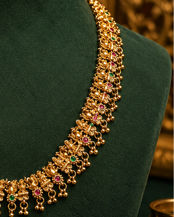 An ornate gold-toned traditional necklace displayed on a dark green velvet jewelry bust, with decorative flowers and warm-toned background elements around it.
