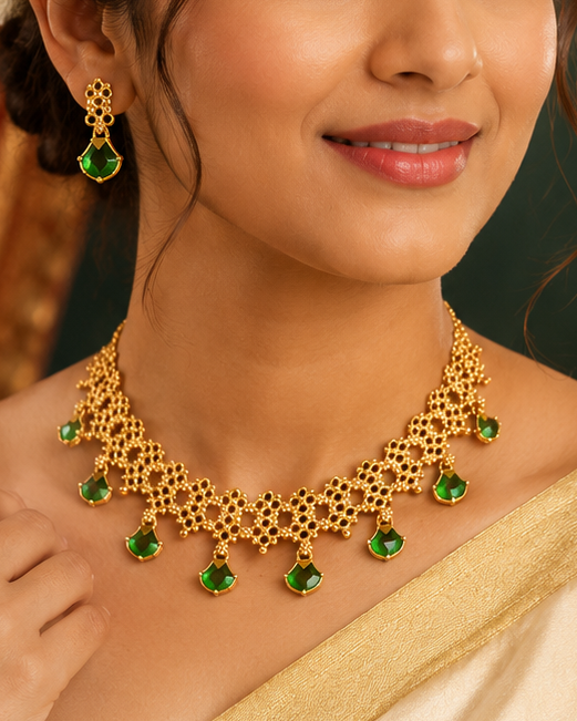 A woman wearing a gold and green traditional necklace and matching earrings, styled with a cream-colored saree, set against a dark green background with a brass lamp.