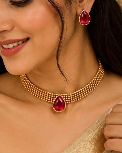 A woman wearing a gold-toned traditional necklace with a red teardrop gemstone and matching red earrings, styled with a cream-colored sari and white hair flowers.
