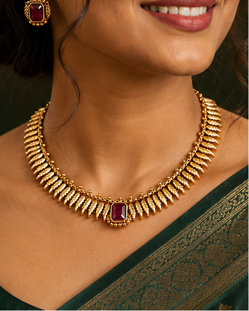 A woman wearing a dark green silk saree with gold zari embroidery and a matching gold necklace with a red stone accent, set against a warm, dark background.