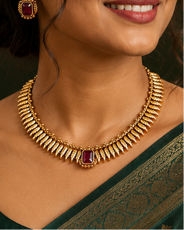 A woman wearing a dark green silk saree with gold zari embroidery and a matching gold necklace with a red stone accent, set against a warm, dark background.