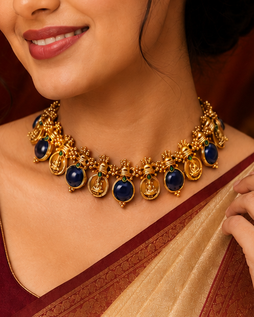 A woman wearing a traditional Indian sari with a deep maroon blouse and a beige sash with gold zari work, styled with a gold and blue stone necklace and a gold bracelet. She is posed against a warm, ornate background with red drapery and a brass pot.