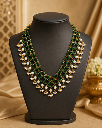 A multi-strand green beaded necklace with gold-toned chains and white pearl-like drops, displayed on a black jewelry bust against a warm beige background with decorative fabric and a small floral vase.