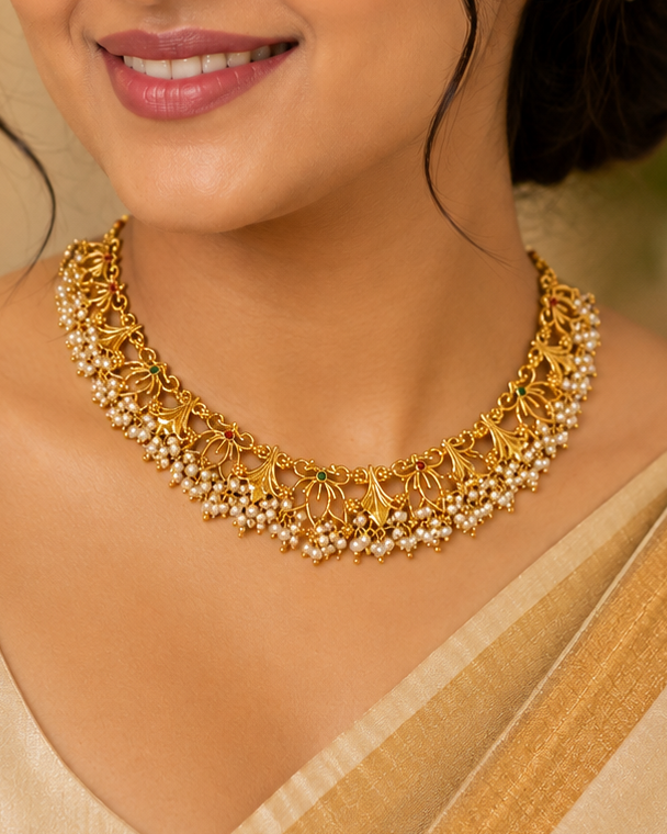A woman wearing a gold traditional necklace with pearl-like accents and a beige saree, shown in a softly lit indoor setting with blurred lights and flowers in the background.