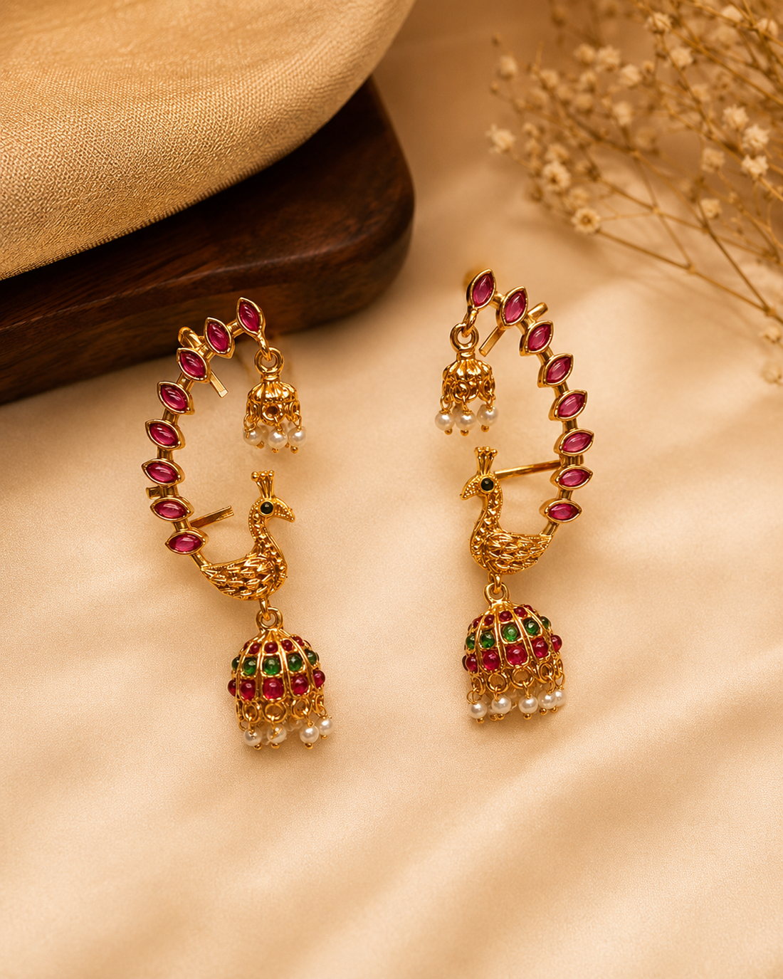 A close-up of a woman wearing ornate gold-toned traditional earrings with red and green stone detailing and small pearl-like accents, shown against a softly blurred warm beige background.