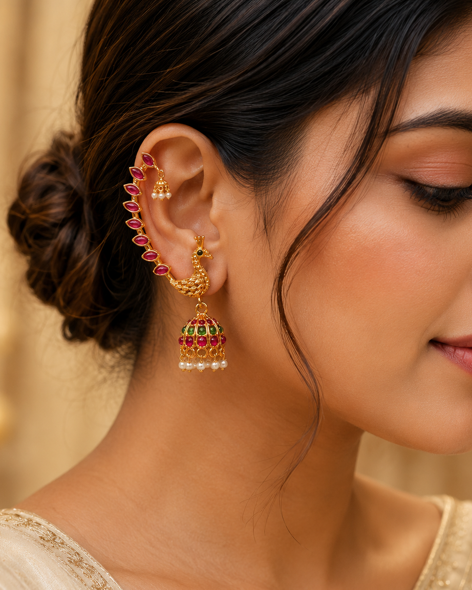 A close-up of a woman wearing ornate gold-toned traditional earrings with red and green stone detailing and small pearl-like accents, shown against a softly blurred warm beige background.
