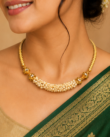 A woman wearing a gold floral choker necklace with small gold and white stones, paired with a green and gold saree. She is shown in a warm, traditional indoor setting with a carved wooden screen in the background.