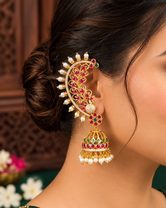 A woman in profile wearing an ornate gold-toned traditional Indian jhumka earring with red stones and white pearl-like accents, paired with a green embroidered blouse.