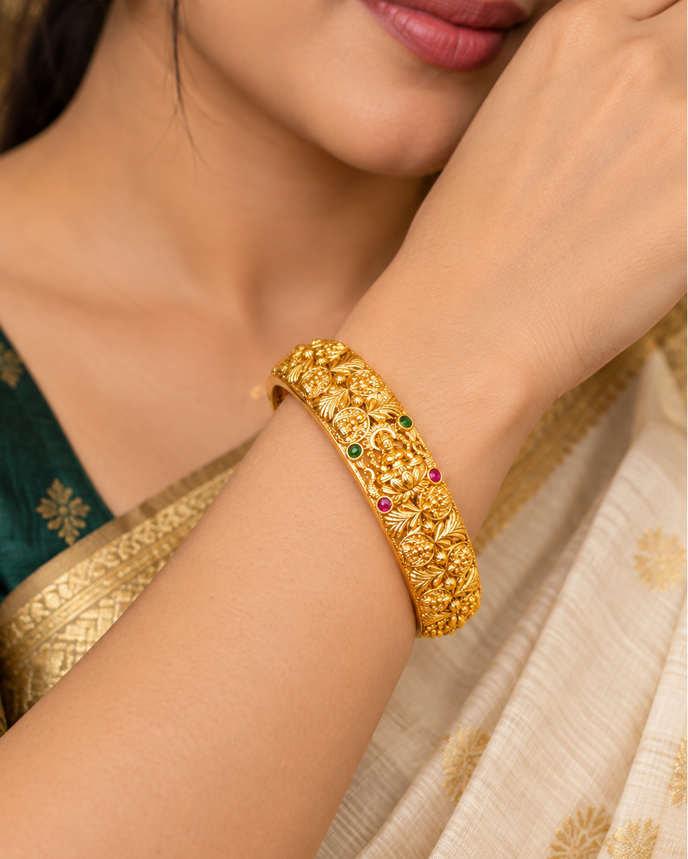 A close-up lifestyle shot of a woman wearing an ornate, gold-plated traditional Indian bangle (kara) decorated with small green and red enamel stones. She is wearing a dark green embroidered blouse and a cream-colored sari.