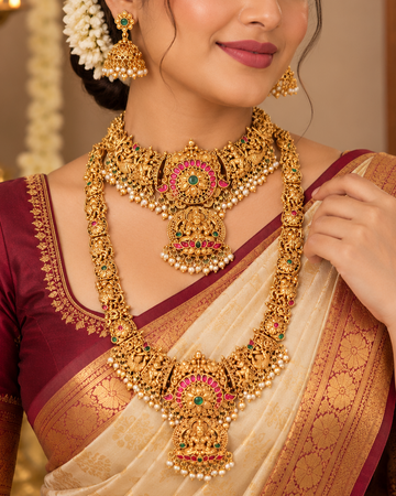 A woman wearing an ornate gold-toned traditional necklace and matching earrings with pink and green stone accents, paired with a cream and maroon sari in an indoor setting.