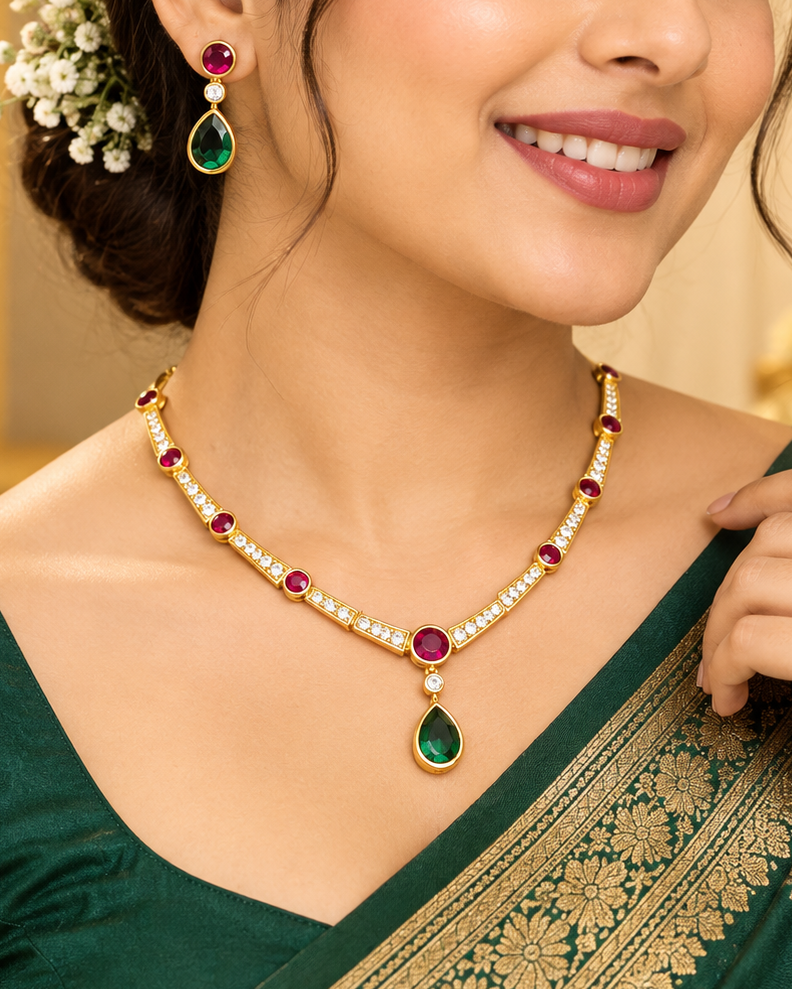 A woman wearing a gold-toned necklace and matching drop earrings with green teardrop stones and red round stones, styled with a dark green saree in an indoor setting.