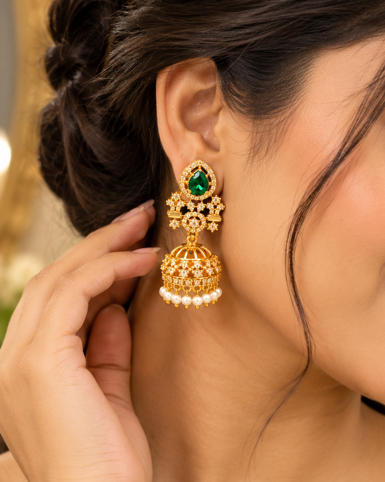 A close-up lifestyle shot of a woman wearing a traditional Indian gold jhumka earring with a green stone and white pearl accents, paired with a dark green saree.
