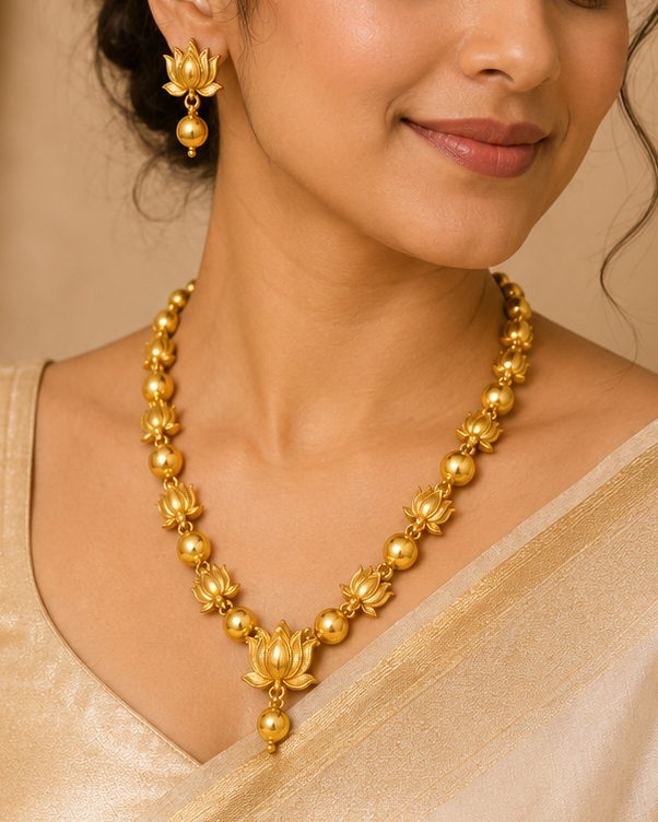 A woman wearing a gold necklace and matching gold earrings, styled with a light beige silk saree, set against a warm beige background with draped curtains.
