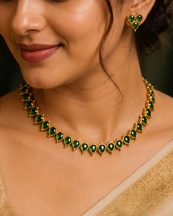 A woman wearing a green and gold necklace and matching earrings, styled with a beige saree in a warm, elegant indoor setting.