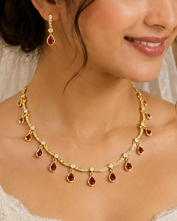 A bride wearing a gold necklace with red teardrop-shaped stones and matching earrings, paired with a white lace wedding dress and holding a bouquet of white roses.