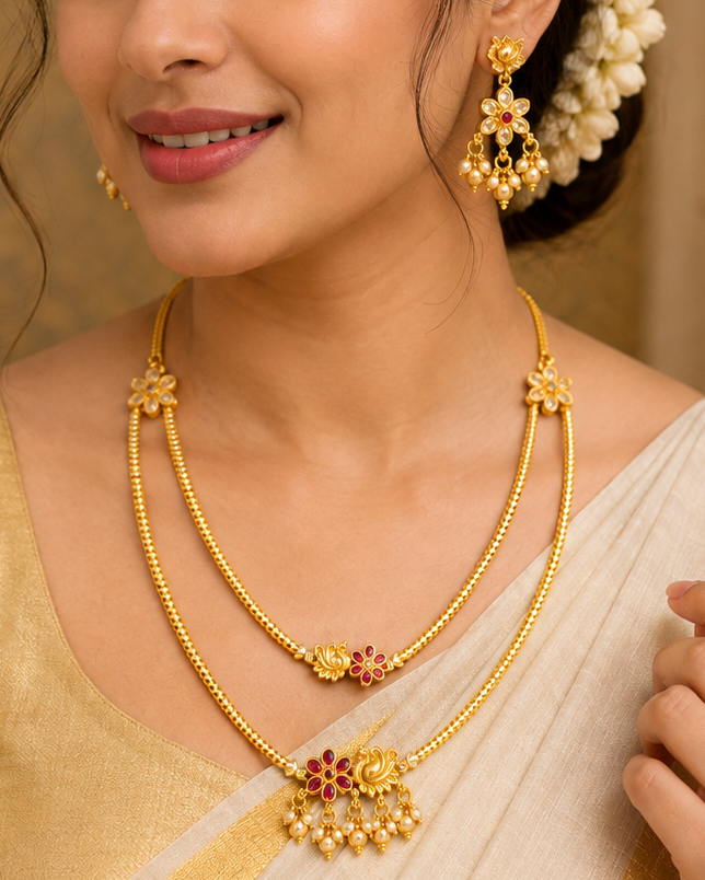 A woman wearing a traditional Indian outfit with a gold and cream saree, styled with a double-layered gold necklace and matching earrings, set against a warm background with lit brass lamps.